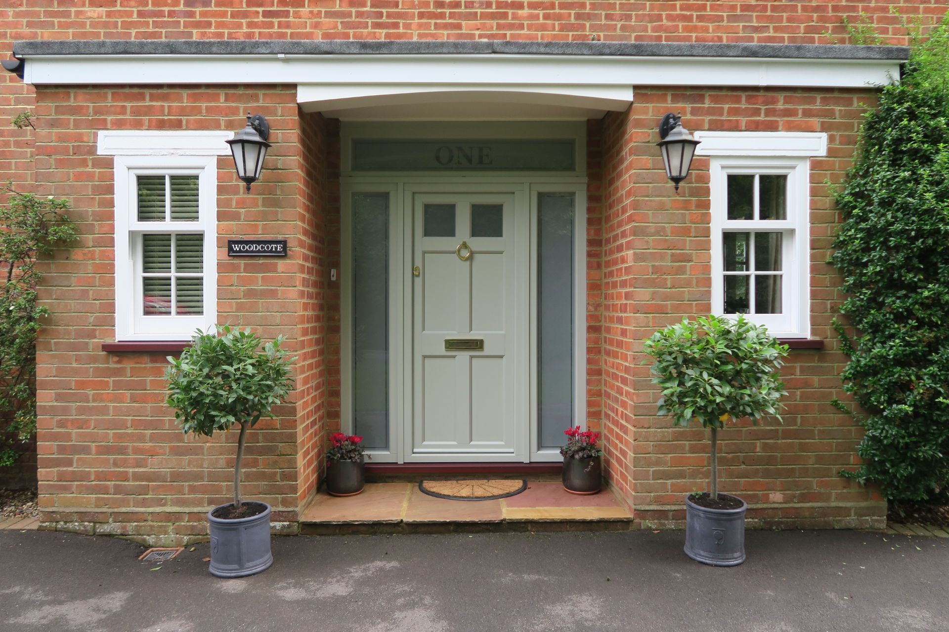 Sage green composite front door with glazed side panels and brass hardware in a brick porch